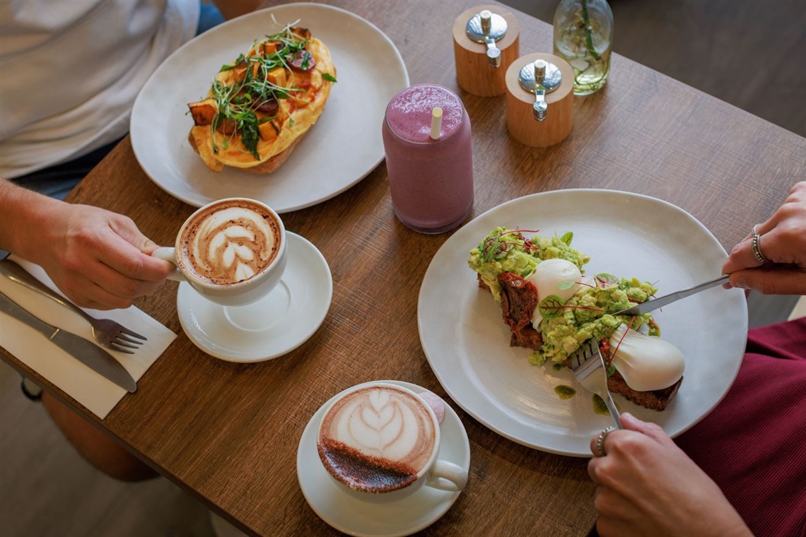 A table with two plates of breakfast food, two coffees, a pink smoothie, salt and pepper and a vase of flowers.