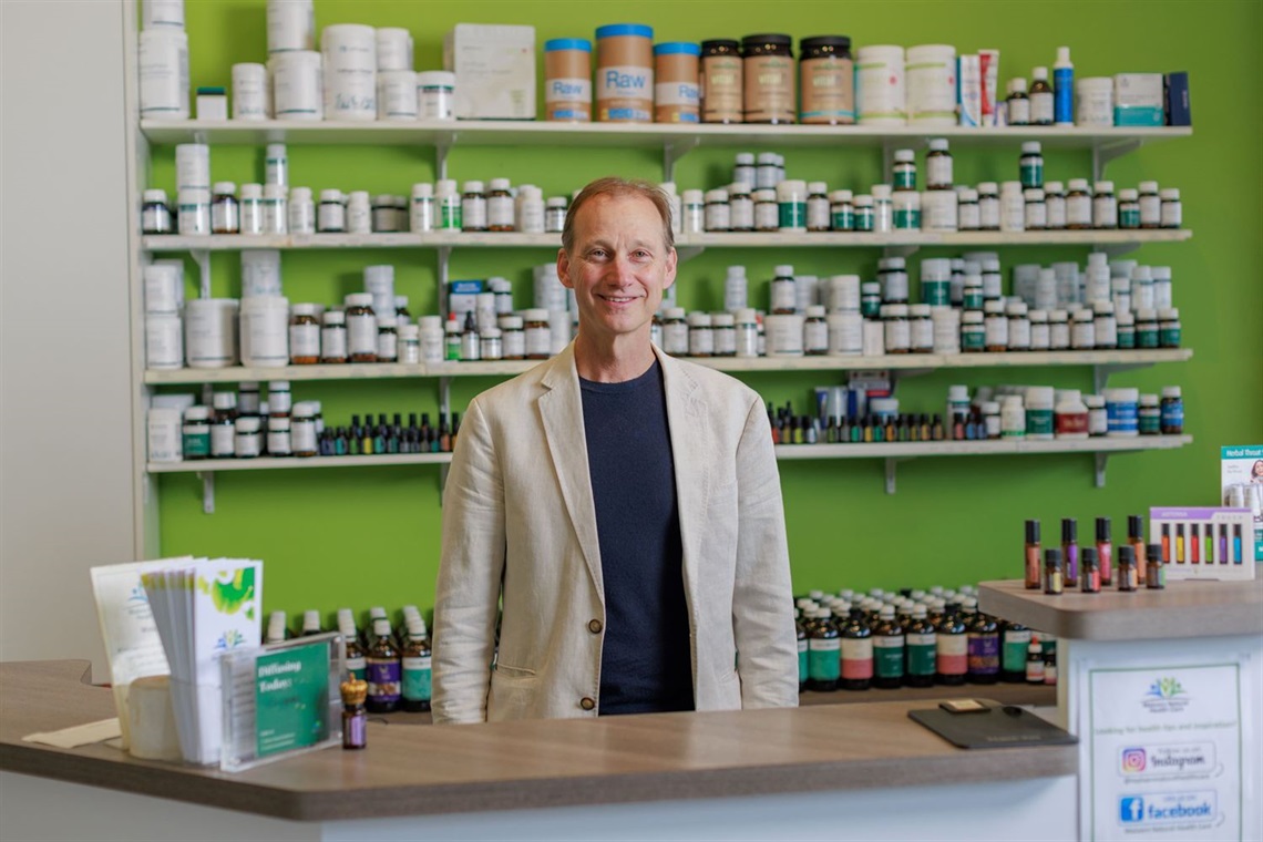 A man in a sports jacket smiling and standing behind a counter with shelves full of medicine, oils and supplements behind him.