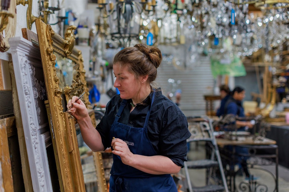 Woman wearing an apron using a paint brush on a gold frame. Chandeliers hang overhead and other people are working in the room behind her.