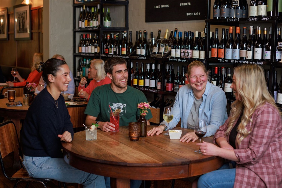 Four young adults sitting around a table smiling at each other. They have an alcoholic drink each before them and shelves of wine bottles behind them.