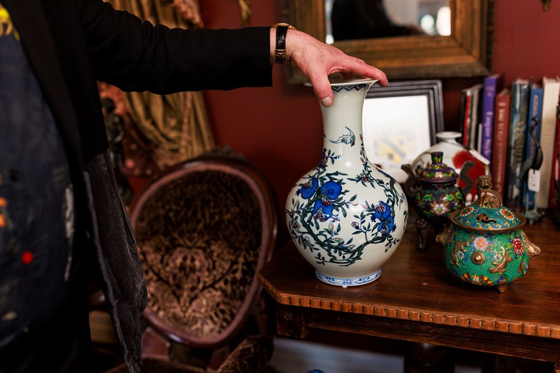 An old wooden table with antique vases, ceramic containers and books. An old mirror is on a wall behind the table. 