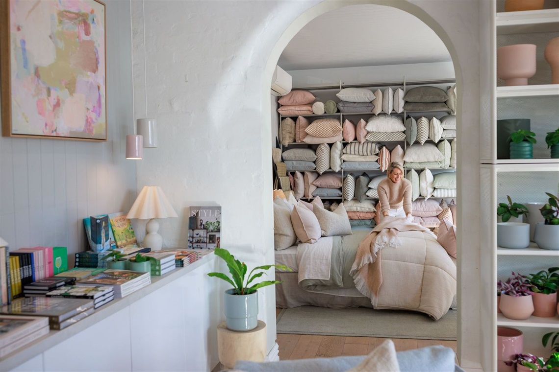 A white-walled shop filled with cushions, books and plants. A smiling woman is styling a bed.