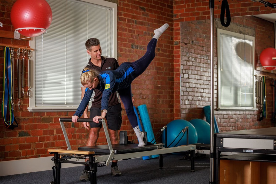 A man assists a woman to achieve correct form as she lifts a leg and supports herself with her hands on a reformer pilates bench. They are in a gym.