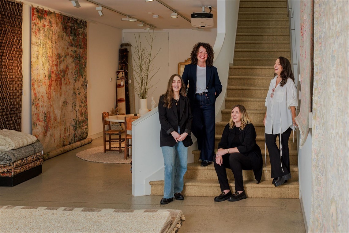 Four smiling women standing at the bottom of a carpetted staircase. There are in a room with rugs on the walls, floor and neatly folded in stacks.