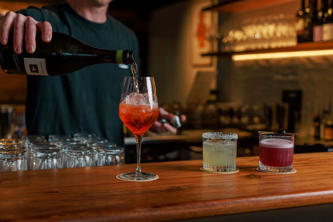 A man pouring wine into a wineglass filled with ice. Two tumblers with cocktails sit on the bar beside the wineglass.