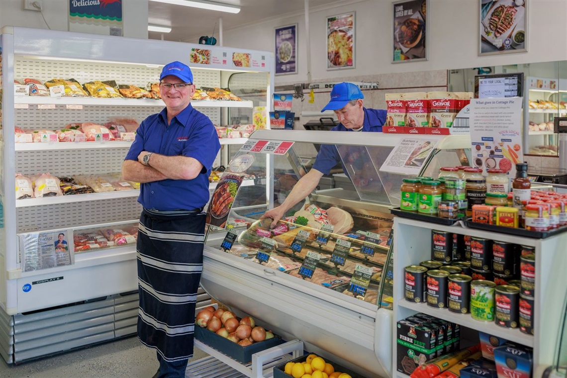 A smiling man in a butcher's apron standing in front of a refrigerated display of meats. There is a shelf stocked with pantry items beside the fridge and some fresh produce.