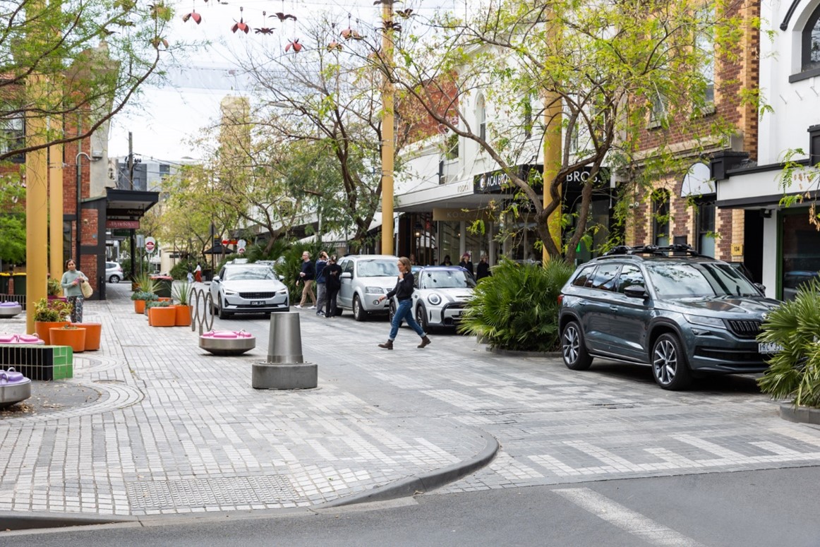 Surburban street with woman crossing road, several parked cars and an approaching car. Tall green trees line the street. There is a wide footpath and speed hump.