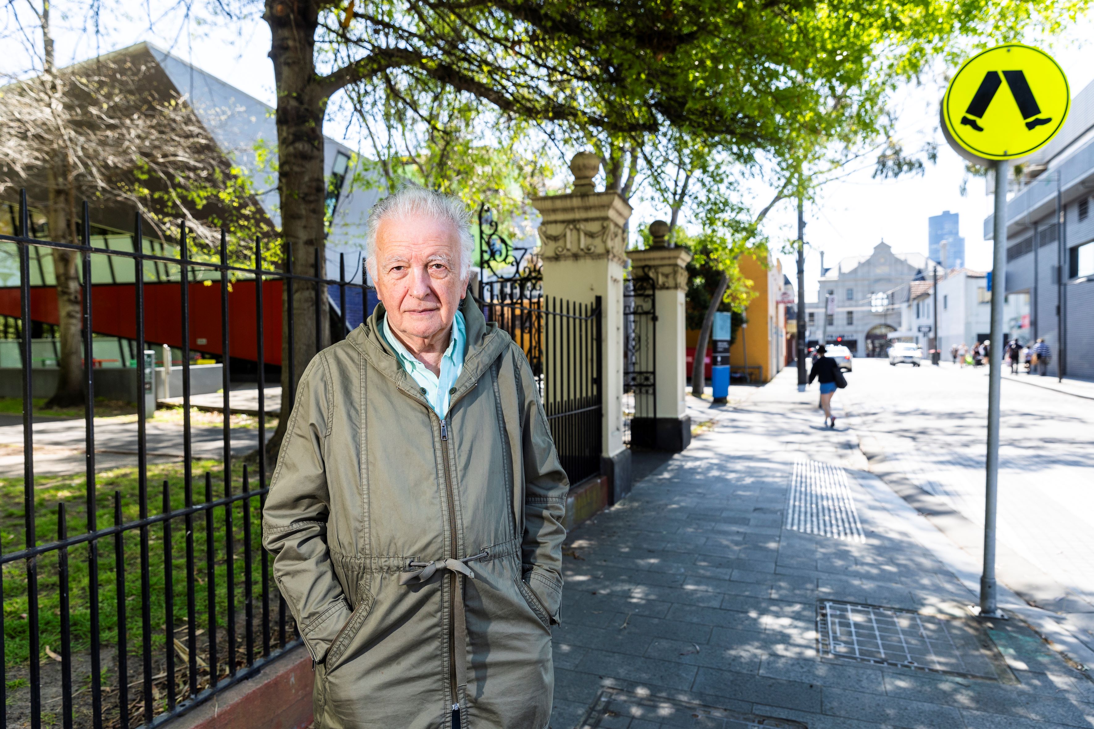 Man standing on suburban footpath with large iron fence and stone entrance in background.