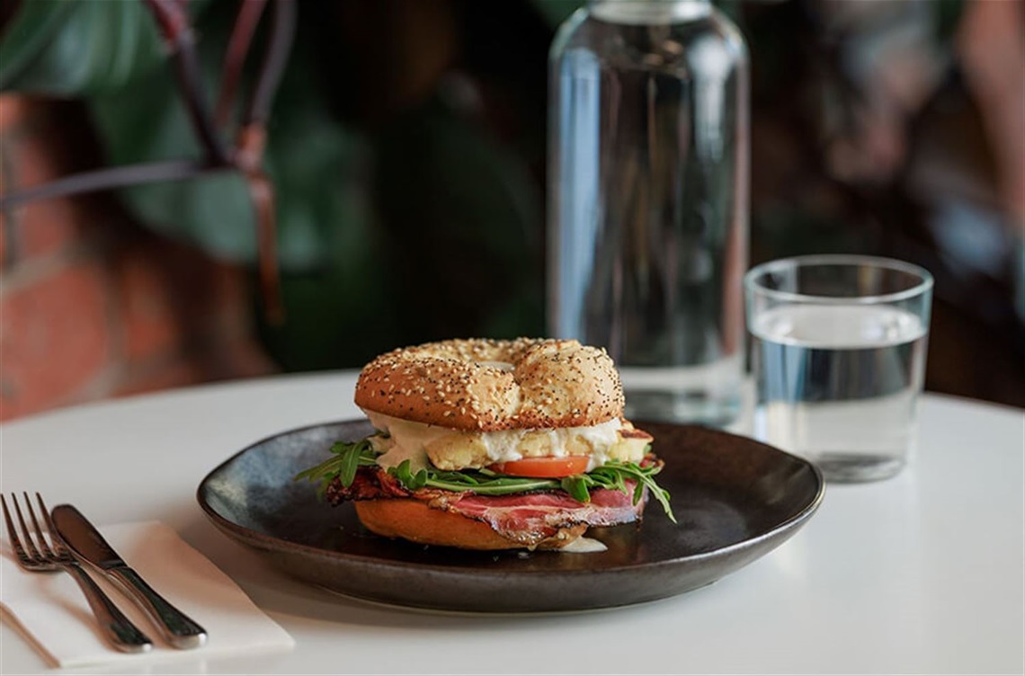 A bagel filled with egg and vegetables sits on a plate at a table alongside cutlery, a napkin, a bottle and a glass of water. An indoor plant grows along the wall behind the table.