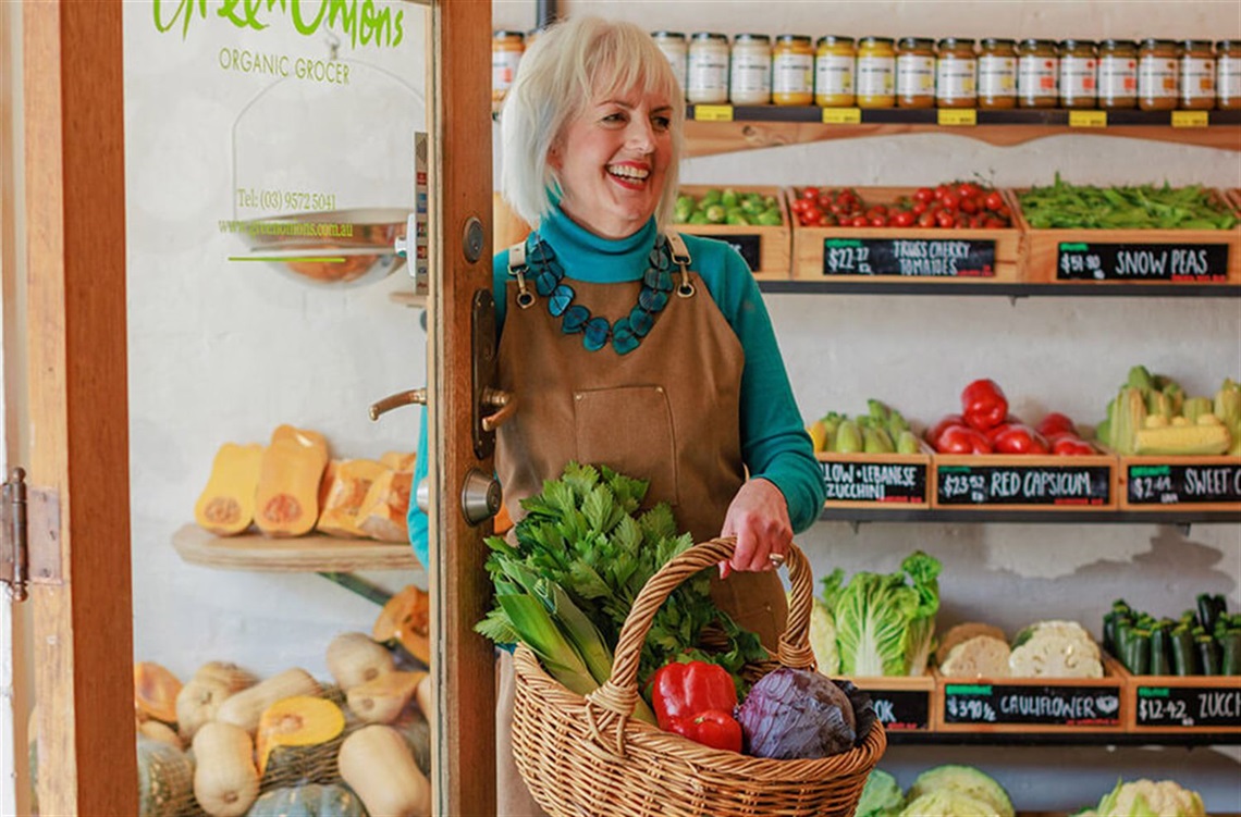 A smiling woman with a basket full of fresh vegetables is standing in a shop with fresh fruit and vegetables. Jars of produce are on shelves.