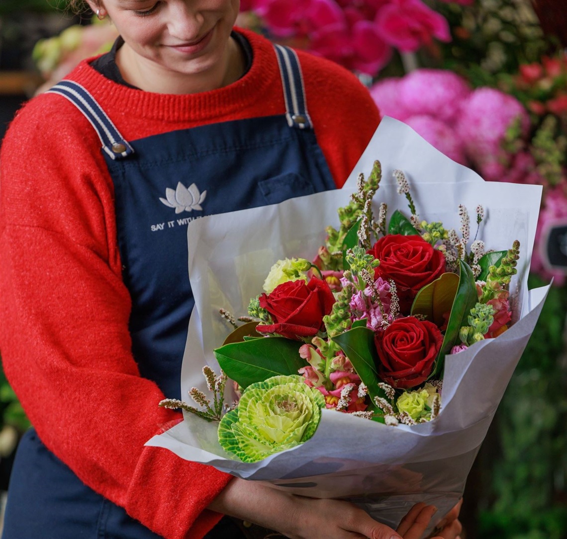 A woman holds a colourful bunch of flowers wrapped in paper. There are flowers behind her.